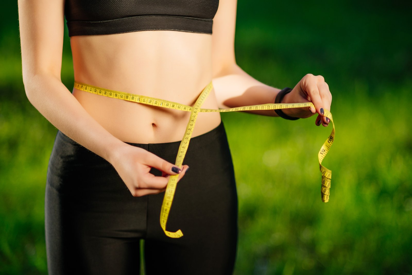 Young woman measuring her thin waist with a tape measure on green nature backgroung, close up. Diet and sports concept
